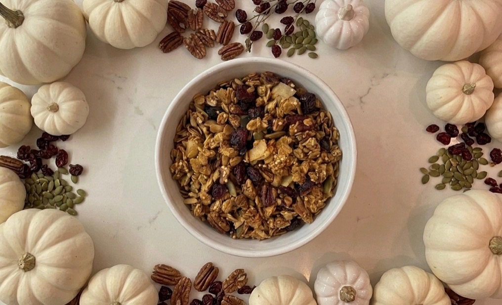 A bowl of Angie's pumpkin spice granola surrounded by white pumpkins, pecans and cranberries